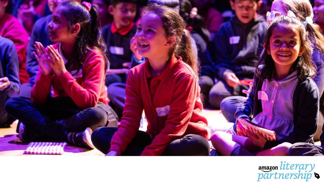 5 or so primary school children in school uniforms are sitting cross-legged on the floor of a dramatically lit auditorium. They are smiling and clutching small goody bags. The Amazon Literary Partnership Logo appears at the bottom right on a white banner that runs underneath the image.