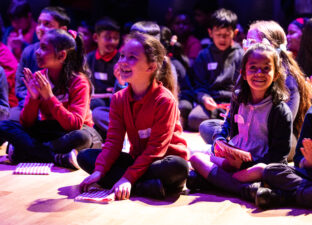 10 or so primary school children in school uniforms are sitting cross-legged on the floor of a dramatically lit auditorium. They are smiling and clutching small goody bags.