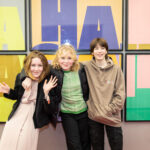 Three young teens stand together in front a of a colourful Ministry of Stories project photo, their arms around each other, smiling