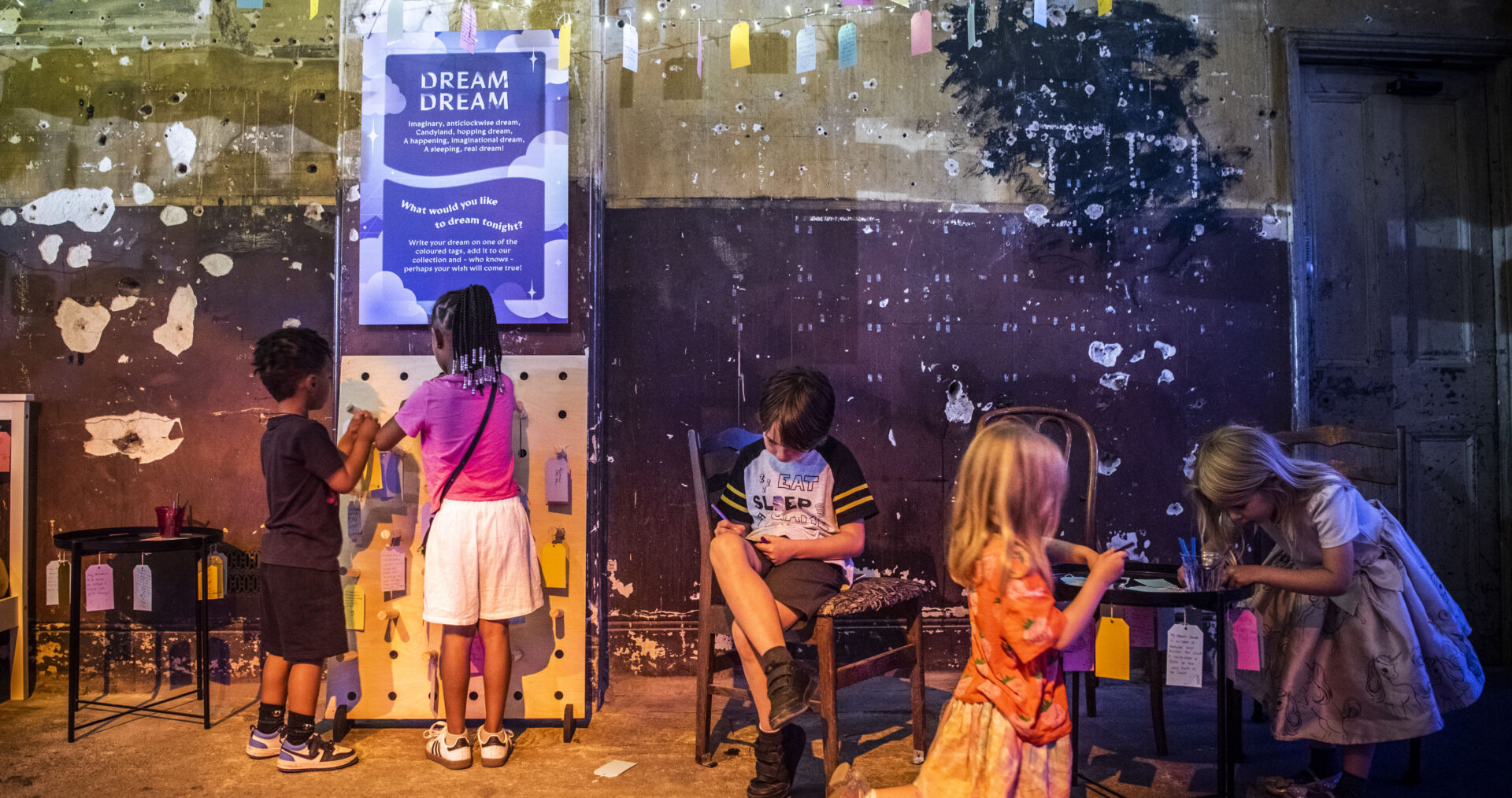 A young boy with short brown hair in shorts and a t-shirt is sitting on a chair and writing on a small colourful paper tag. Four other children are also in the picture, either writing on tags or hanging up their tags on a board. They are in the Longing Room at DreamState, a multi-sensory exhibition of young people's writing.