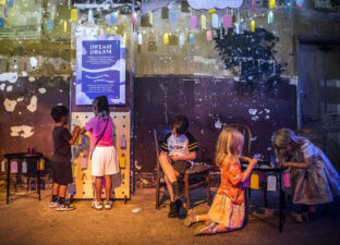 A young boy with short brown hair in shorts and a t-shirt is sitting on a chair and writing on a small colourful paper tag. Four other children are also in the picture, either writing on tags or hanging up their tags on a board. They are in the Longing Room at DreamState, a multi-sensory exhibition of young people's writing.