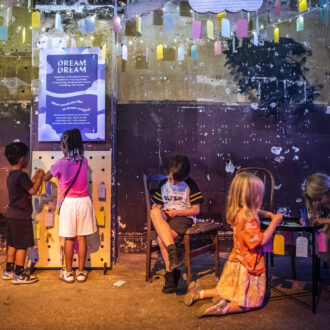 A young boy with short brown hair in shorts and a t-shirt is sitting on a chair and writing on a small colourful paper tag. Four other children are also in the picture, either writing on tags or hanging up their tags on a board. They are in the Longing Room at DreamState, a multi-sensory exhibition of young people's writing.