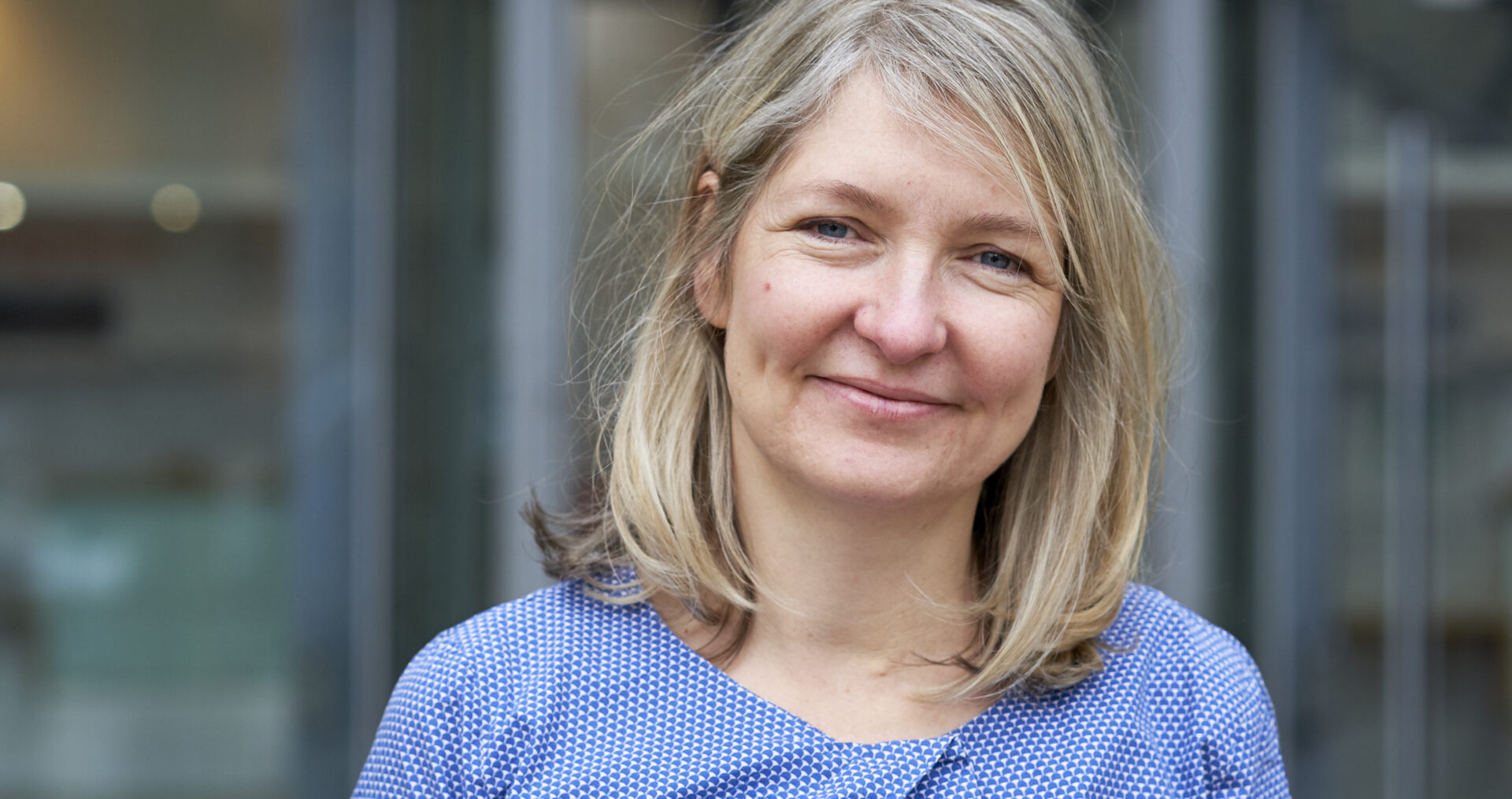 Lucy, a white woman with shoulder length blonde hair, stands smiling at the camera. She wears a light blue, long sleeve top and stands in front of the Hackney Council buidling.