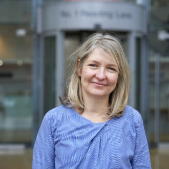 Lucy, a white woman with shoulder length blonde hair, stands smiling at the camera. She wears a light blue, long sleeve top and stands in front of the Hackney Council buidling.