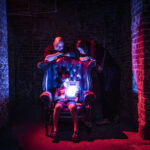A child sits in a big leather chair, flanked by her parents, reading a scary story by flashlight in a dark lit room
