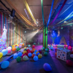 A big room is filled sith party streamers, disco lights and bright colourful baloons. There is also a giant, spotty cardboard leopard that sits to one side against a brick wall.