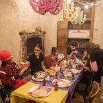 A group of people sit around a dining table, in a brick, desolate basement space, laughing and enjoying the colourful plates and writing laid out underneath colourful paper chandeliers.