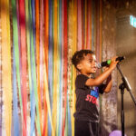 A young boy, standing in front of colourful streamers, sings into a microphone looking joyful.
