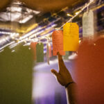 A hand reaches up to colourful luggage tags hanging from the ceiling. They are covered in writing showing different people’s dreams including owning 500 hamsters.