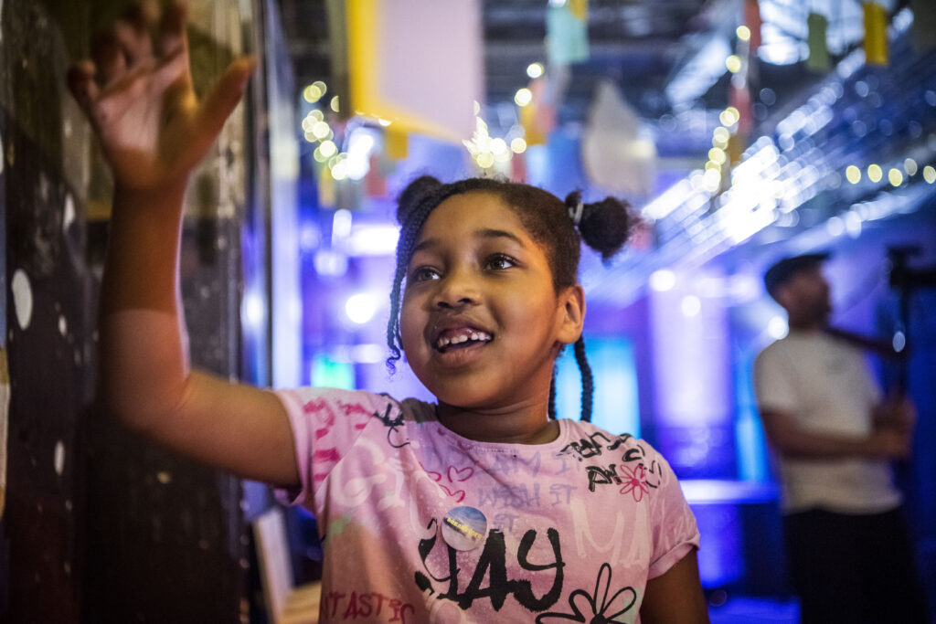 A young girl, in a pink t-shirt and space buns - smiles as she reads some writing on the wall