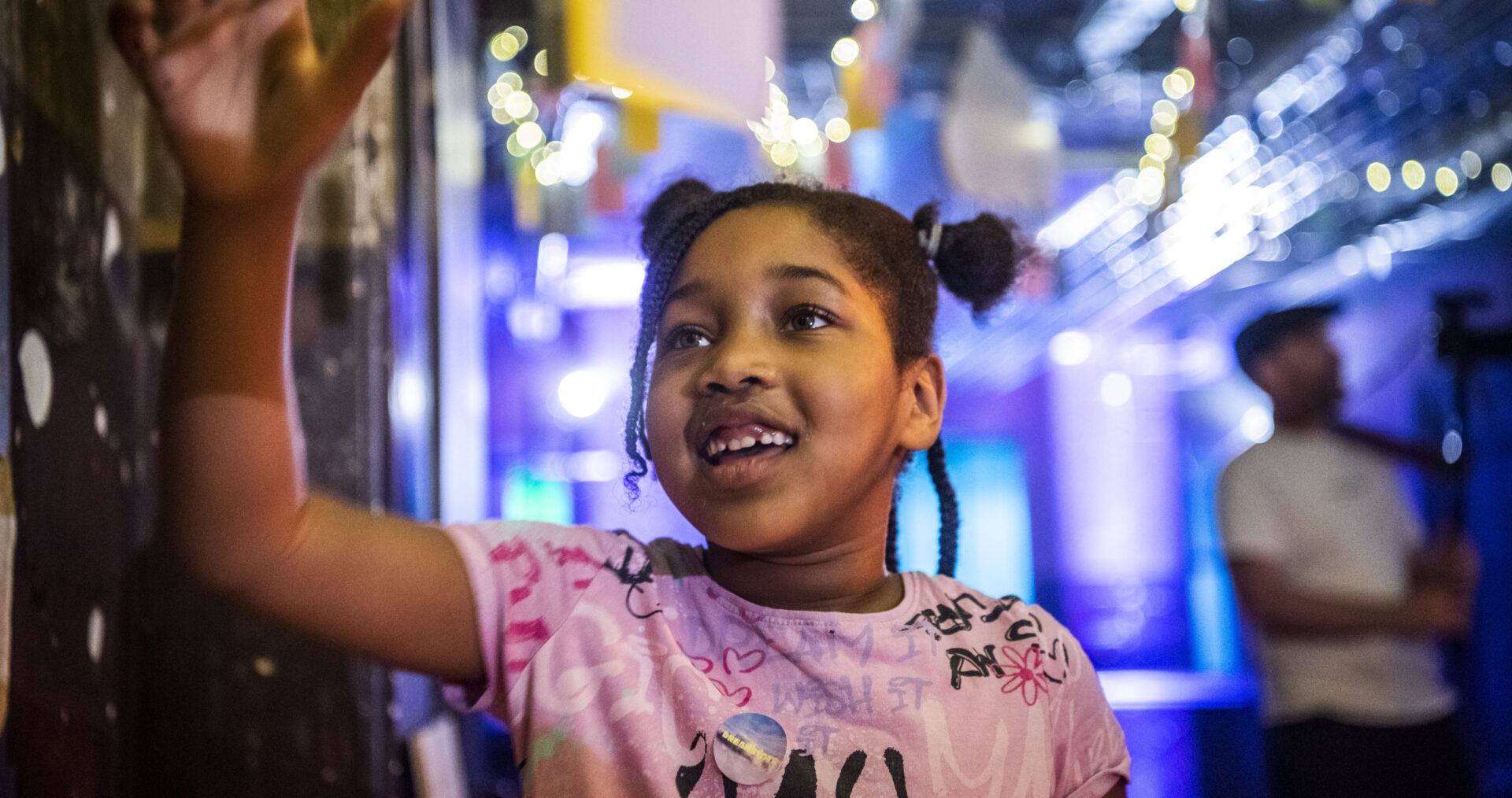 A young girl, in a pink t-shirt and space buns - smiles as she reads some writing on the wall