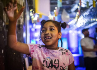 A young girl, in a pink t-shirt and space buns - smiles as she reads some writing on the wall