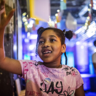 A young girl, in a pink t-shirt and space buns - smiles as she reads some writing on the wall