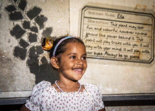 A young girl in a white dress and space buns in her hair, smiles broadly standing in front of a wall which has pictures of plants and writing projected onto it