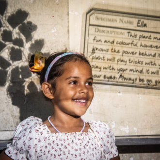 A young girl in a white dress and space buns in her hair, smiles broadly standing in front of a wall which has pictures of plants and writing projected onto it