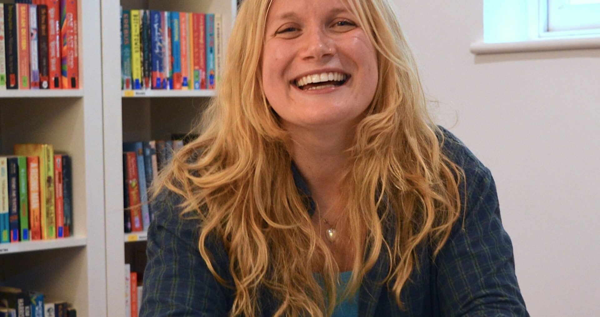 Sarah is a white woman with long blonde hair, down past her shoulders. She is beaming at the camera with her hands clasped on a table in front of her. She is wearing a dark blue check shirt with long sleeves.