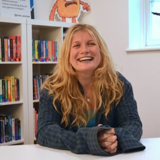 Sarah is a white woman with long blonde hair, down past her shoulders. She is beaming at the camera with her hands clasped on a table in front of her. She is wearing a dark blue check shirt with long sleeves.