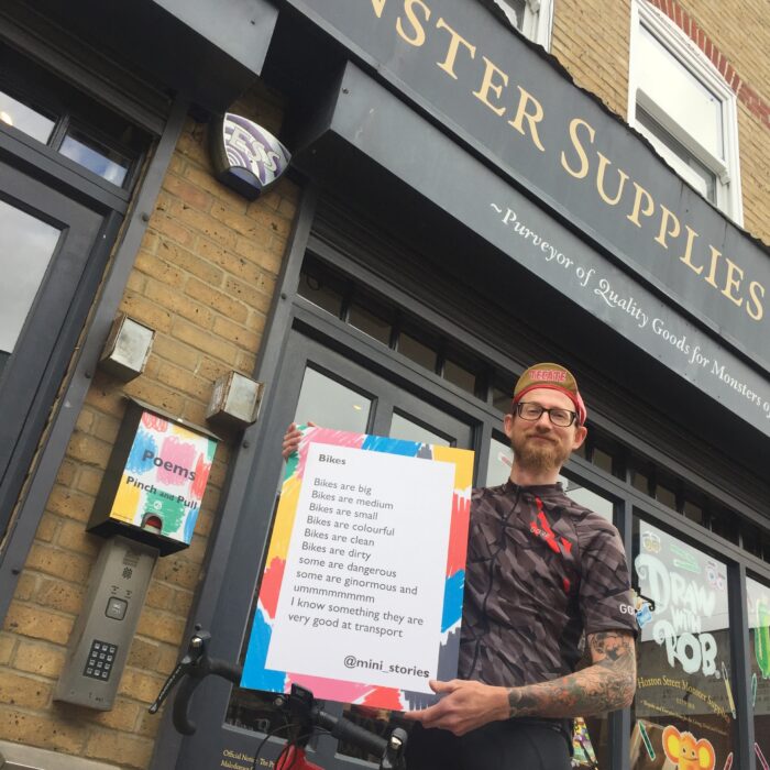Greg - a slim white man who wears glasses and has a brown beard - is wearing a cycling jersey and cap and holding up a poem about Bikes by one of our young writers. He is standing in front of Hoxton Street Monster Supplies shopfront and his road bike is leaning against him.