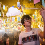 A young teenage boy with glasses and a white t-shirt reads writing on a luggage tag, hung from the ceiling, at DreamState a Ministry of Stories exhibition.