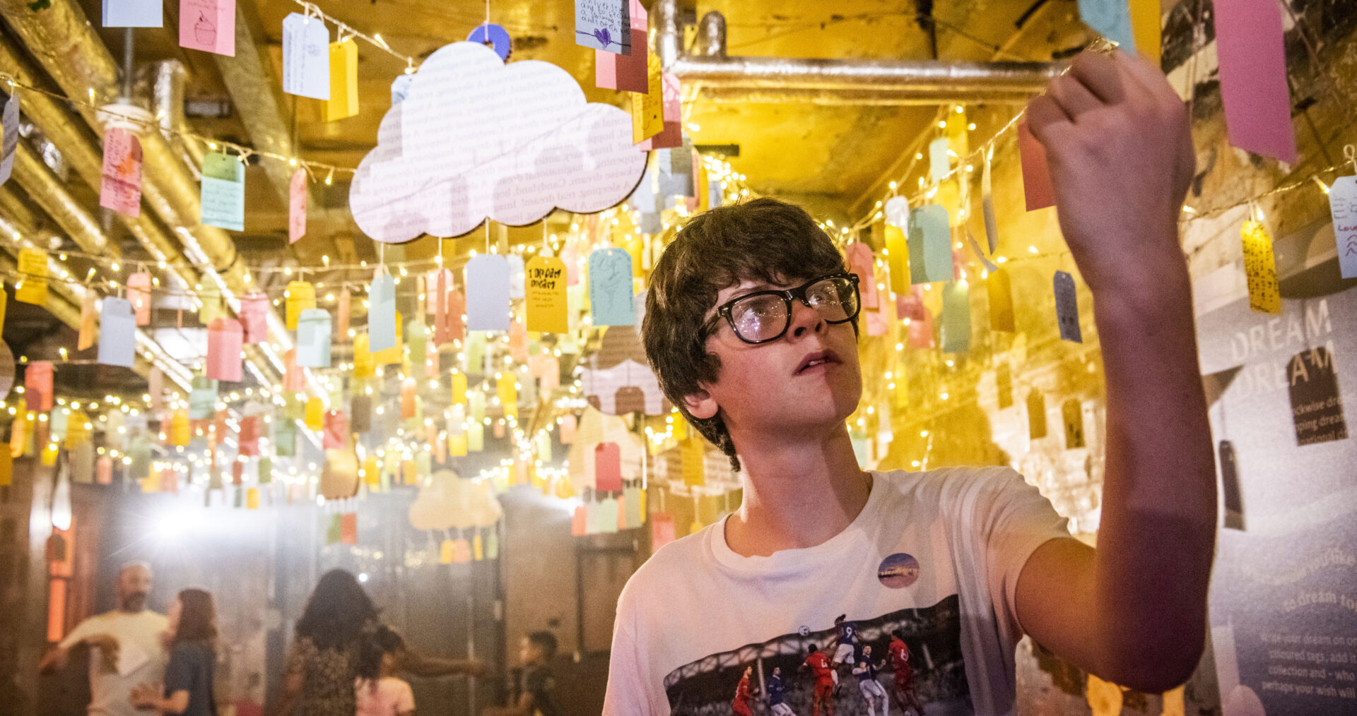 A young teenage boy with glasses and a white t-shirt reads writing on a luggage tag, hung from the ceiling, at DreamState a Ministry of Stories exhibition.