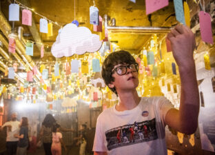 A young teenage boy with glasses and a white t-shirt reads writing on a luggage tag, hung from the ceiling, at DreamState a Ministry of Stories exhibition.
