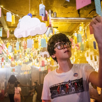 A young teenage boy with glasses and a white t-shirt reads writing on a luggage tag, hung from the ceiling, at DreamState a Ministry of Stories exhibition.