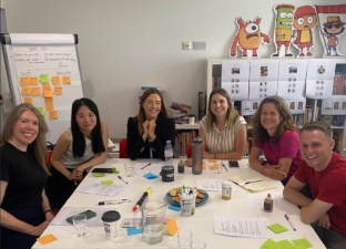 A team of 5 people are gathered around a table at Ministry of Stories HQ brainstorming. There are pens, papers and post-its spread out on the table and everyone is smiling at the camera.