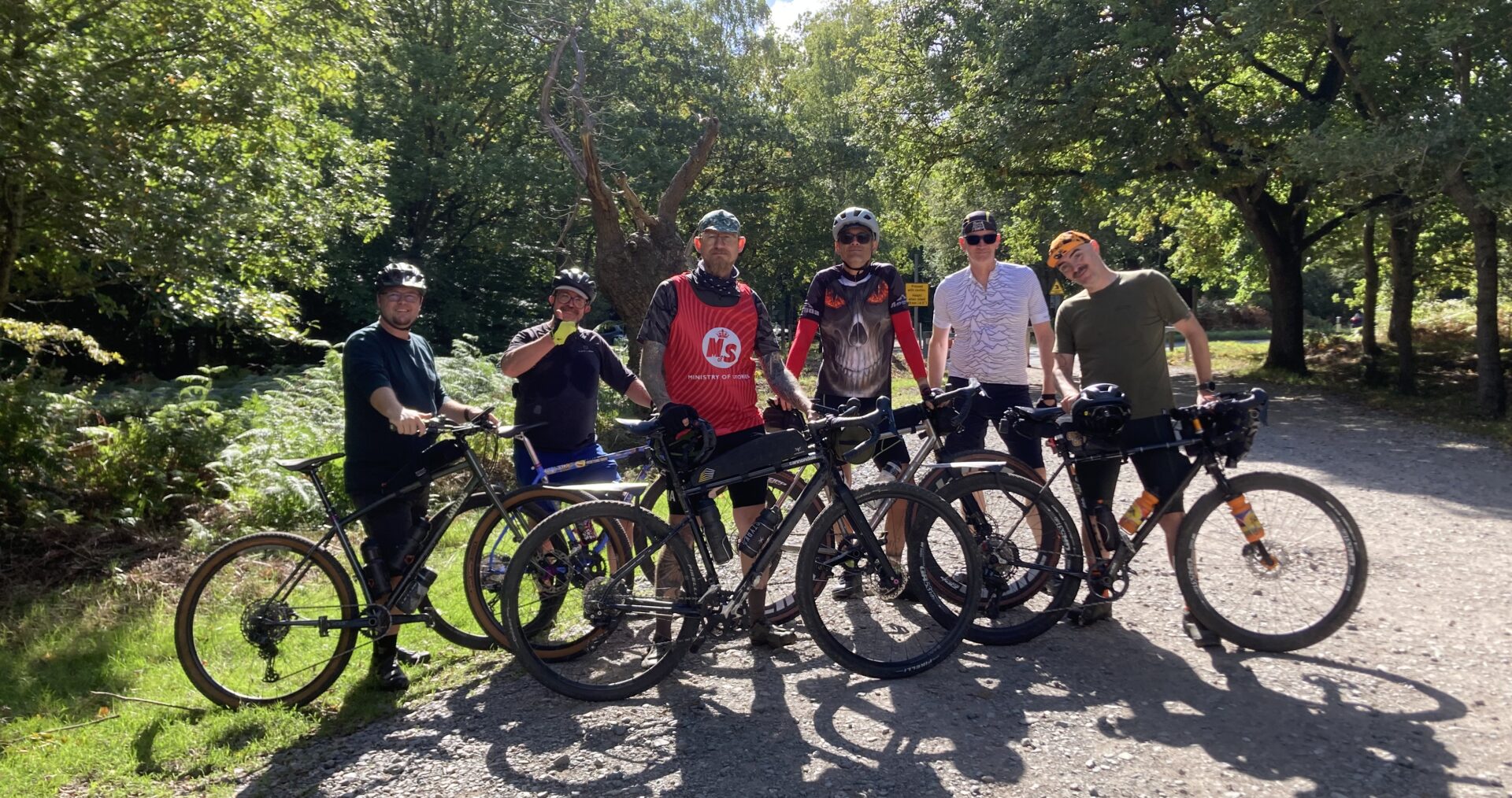 A group of six men in athletic clothing pose with their bikes in the forest. They are on a dirt path and behind them are trees and a blue sky with some clouds.