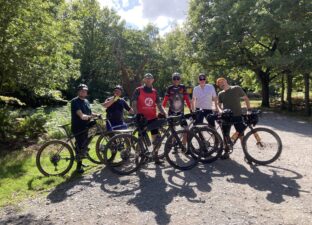 A group of six men in athletic clothing pose with their bikes in the forest. They are on a dirt path and behind them are trees and a blue sky with some clouds.