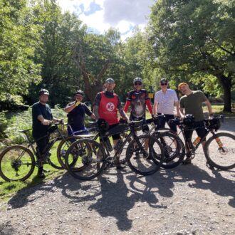 A group of six men in athletic clothing pose with their bikes in the forest. They are on a dirt path and behind them are trees and a blue sky with some clouds.