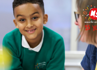 A young boy, with shore dark hair and green jumper, leans in and smiles broadly at the camera. Beside him sits a happy writing mentor, as they write together at a white table.