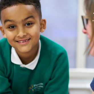A young boy, with shore dark hair and green jumper, leans in and smiles broadly at the camera. Beside him sits a happy writing mentor, as they write together at a white table.