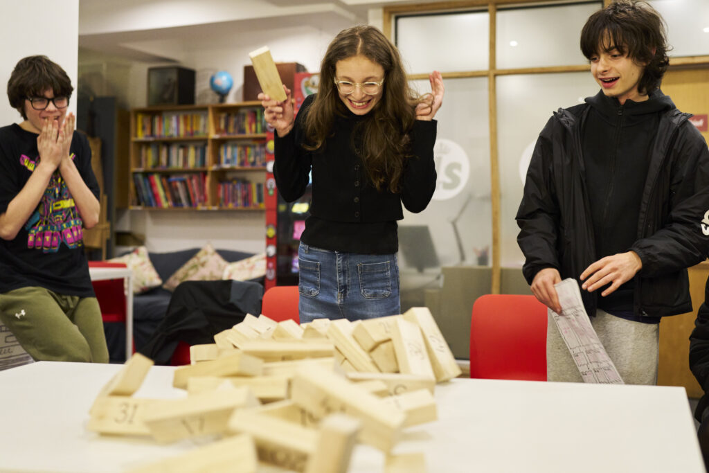 Three teenage writers stand around a recently-fallen Jenga tower. They are smiling and reacting.