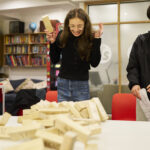 Three teenage writers stand around a recently-fallen Jenga tower. They are smiling and reacting.