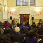A crowd of people are sitting and watching a young person give a speech. This is at the Speak Up sharing event, where young people from Bridge Academy are giving speeches they have written about topics close to their heart.