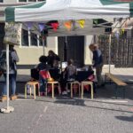 Two young people are sitting and writing at a market stall with bunting. They are taking part in the Fun Palace workshop - Make your own Monster Trading Card.