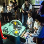 A group of children and adults explore the Control Room at the exhibition DreamState. They are looking at a colourful machine with pipes leading out of it - the Dream Machine.