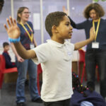 A young writer stands in Ministry HQ with his arms out, playing a game.