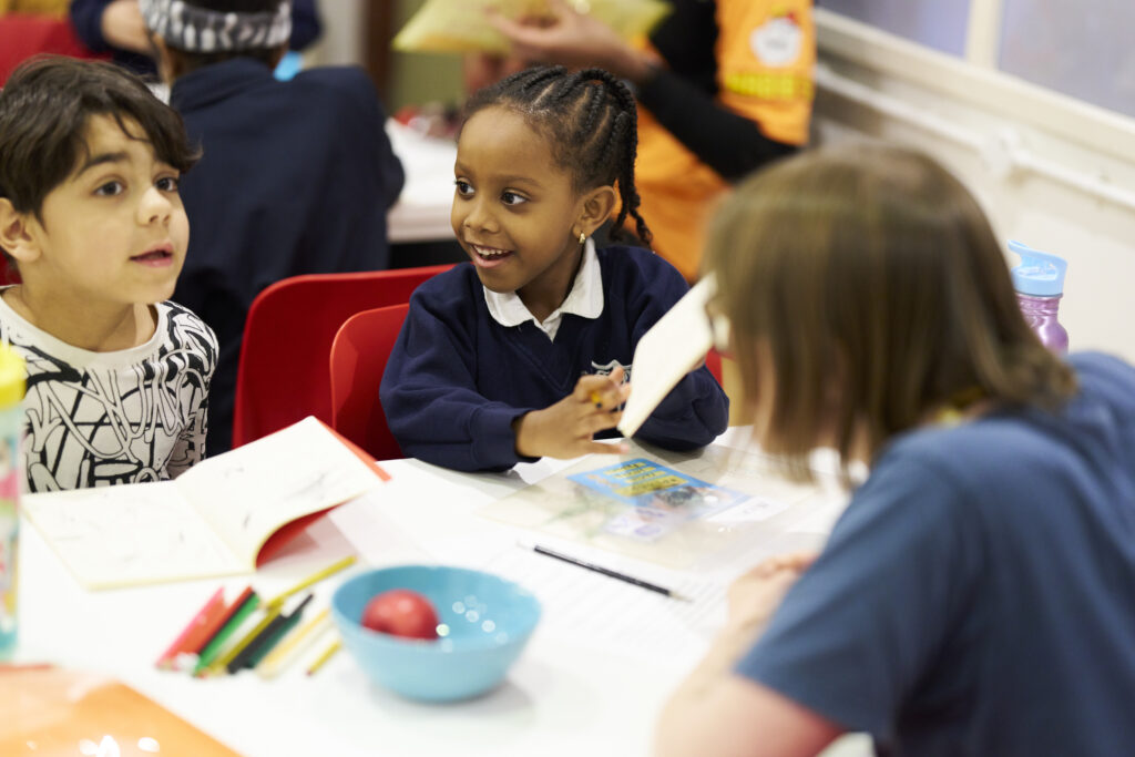 Two young writers and a writing mentor sit around a table at our writing centre, discussing ideas. They are smiling and chatting.