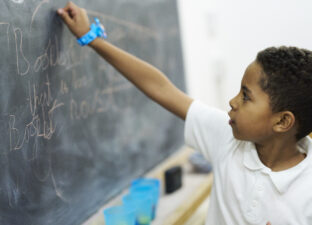 A young writer in a white polo shirt writes with chalk on the blackboard at our writing centre. He is wearing a blue watch and concentrating on his writing.