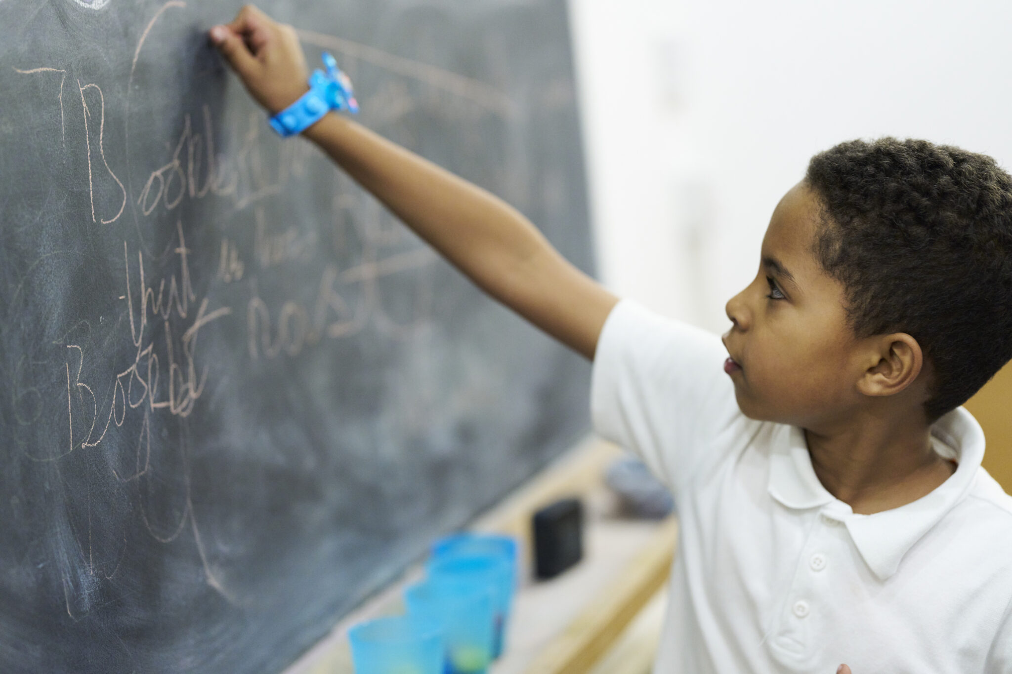 A young writer in a white polo shirt writes with chalk on the blackboard at our writing centre. He is wearing a blue watch and concentrating on his writing.