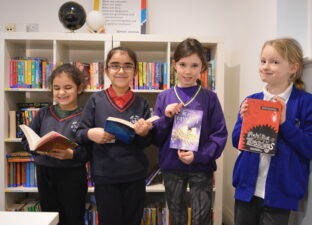 Four young girls are standing in a relaxed line together all holding up books they enjoy reading, towards the camera.
