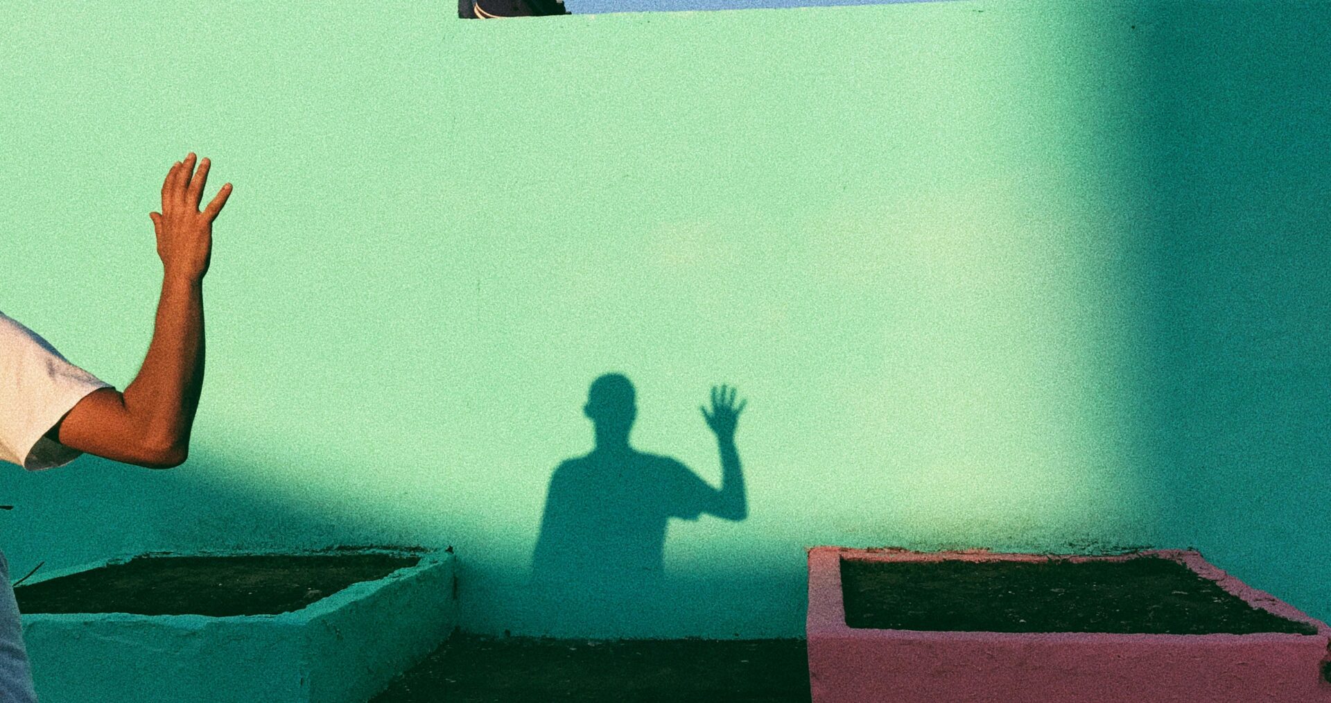 A hand appears from off camera on the left, and creates a shadow in the distance - against a green wall - of a man waving. Thy sky is blue and the bright and sunny.