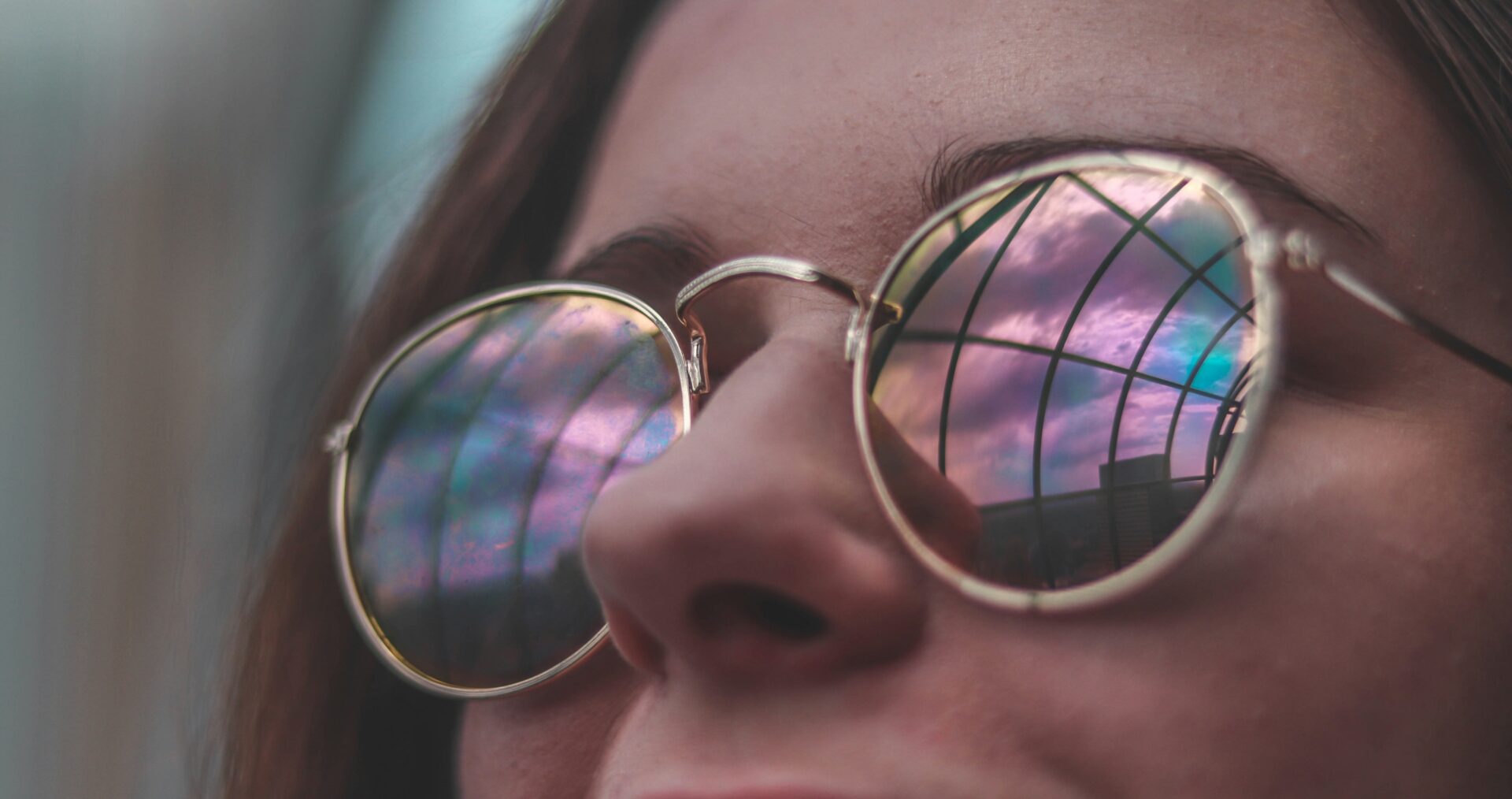 A young woman with dark brown hair and rainbow coloured sunglasses smiles at someone or something out of shot.