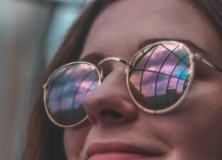 A young woman with dark brown hair and rainbow coloured sunglasses smiles at someone or something out of shot.