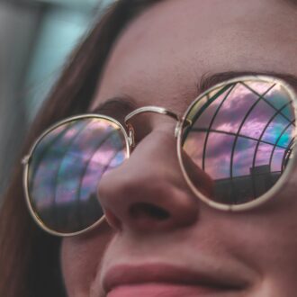 A young woman with dark brown hair and rainbow coloured sunglasses smiles at someone or something out of shot.