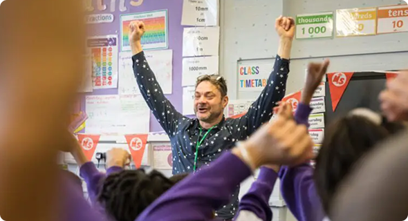 A male teacher - with short dark hair, wearing glasses on his head and bright purple shirt - stands at the front of a classroom with his his arms raised in excitement and the class respond, cheering.