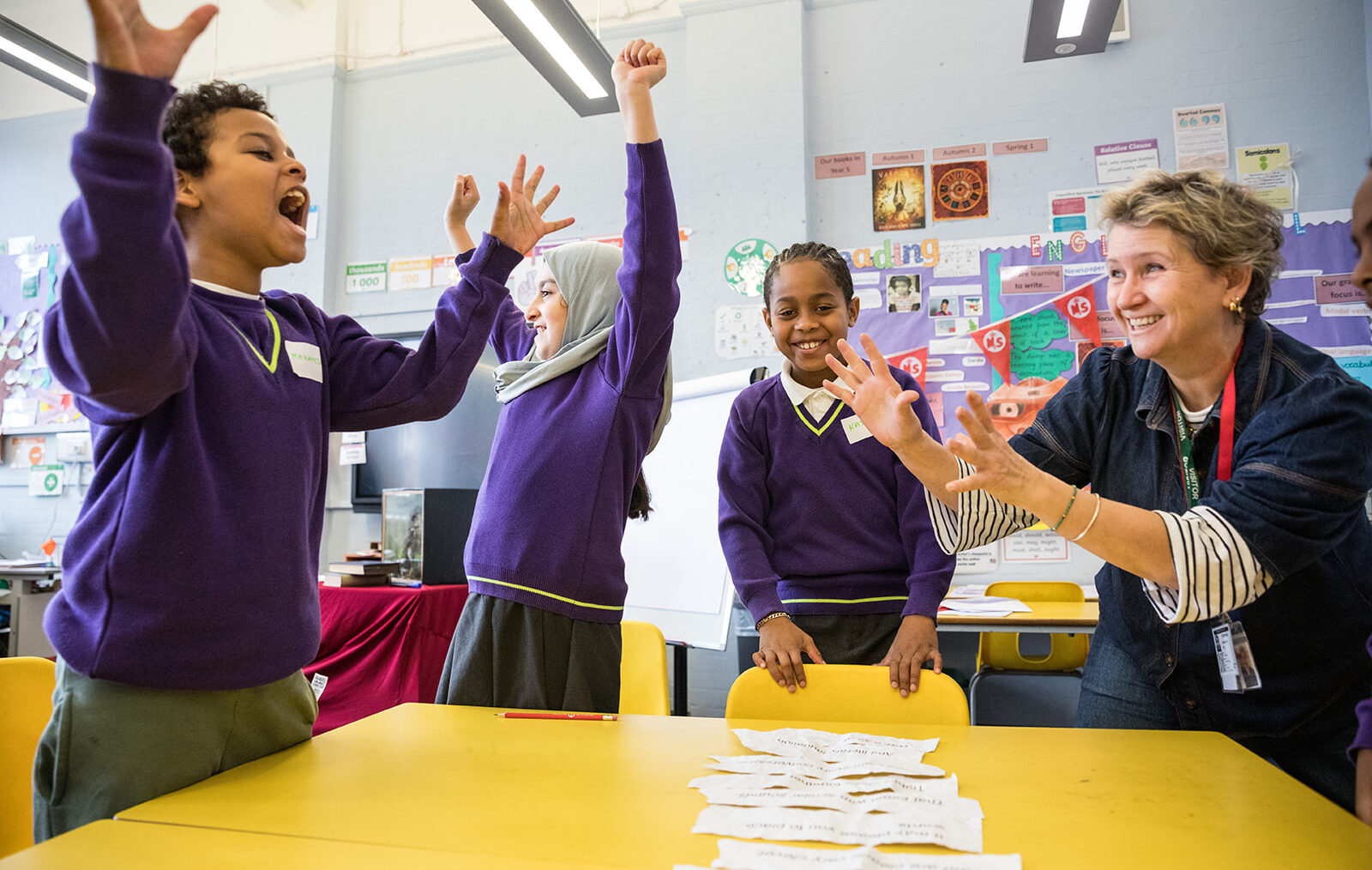 Three primary aged children celerate with big smiles and arms in the air as they stand next to a Ministry writing mentor and a yellow school table