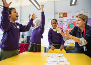 Three primary aged children celerate with big smiles and arms in the air as they stand next to a Ministry writing mentor and a yellow school table