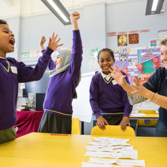 Three primary aged children celerate with big smiles and arms in the air as they stand next to a Ministry writing mentor and a yellow school table